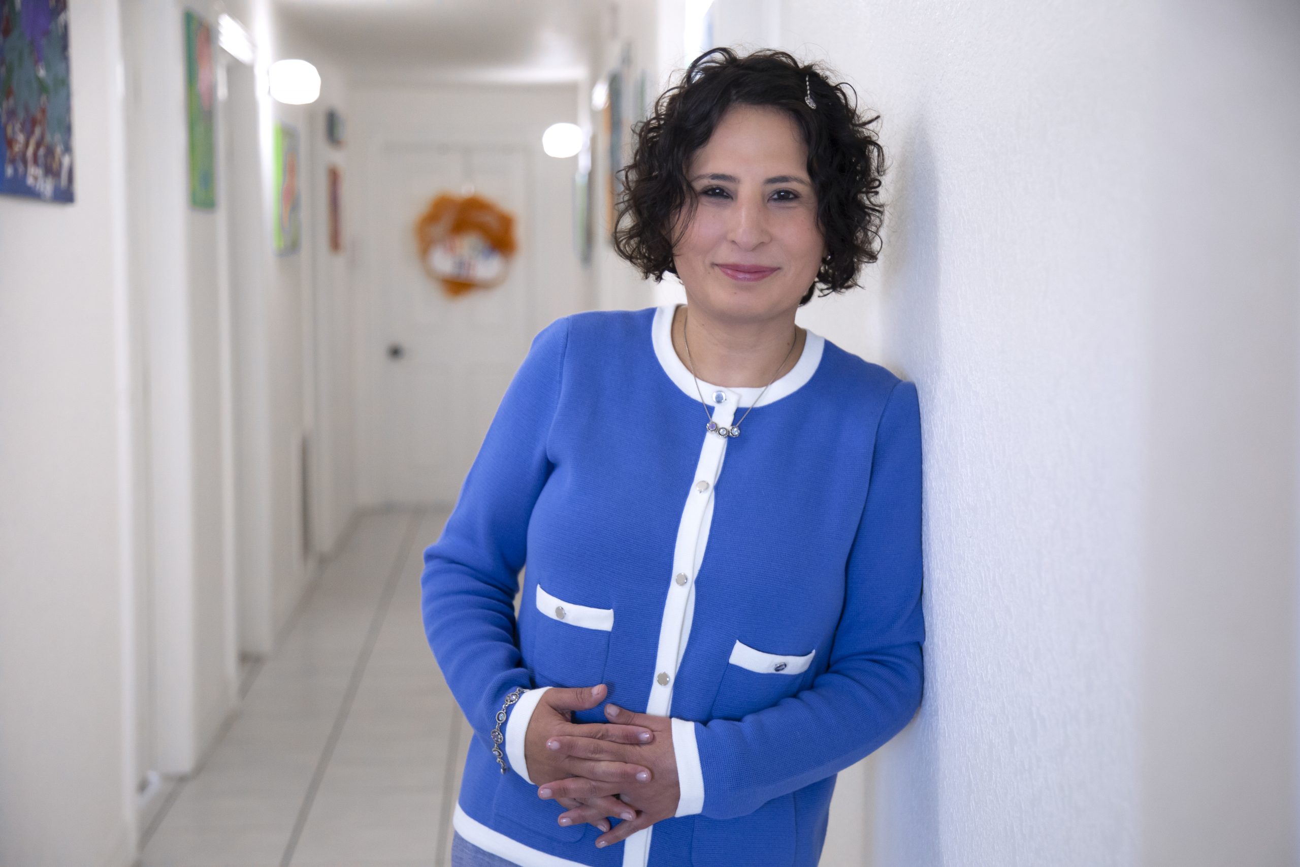 Photograph of Gaby Garcia in the offices of PSP Counseling and its nonprofit sister organization, Dream Big. Garcia, wearing a royal blue sweater with white piping, leans her left shoulder against a wall as she looks toward the camera.