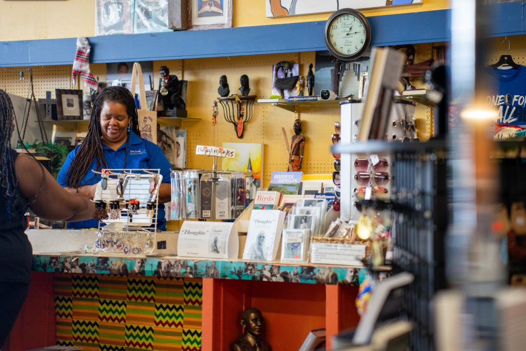 A photograph of a person assisting a customer at a Fort Worth bookshop. A clock hangs on the wall, and small products and trinkets decorate the shelves in the foreground and background.