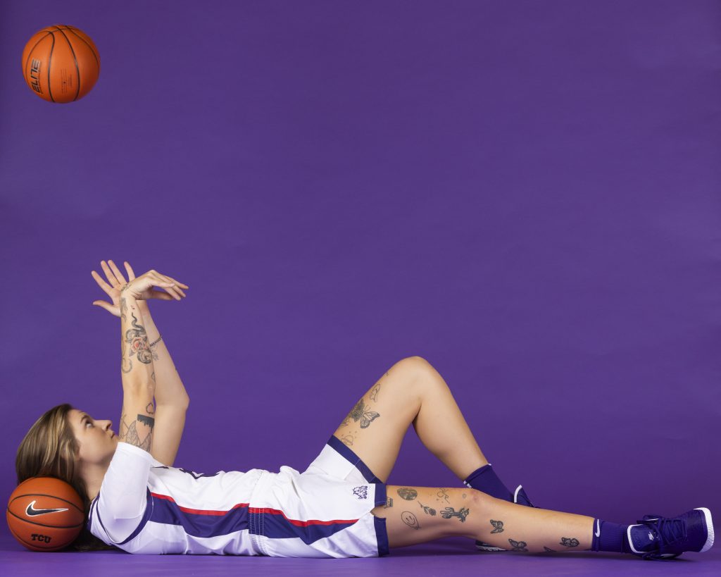 Photograph showing TCU women’s basketball player Sedona Prince laying on her back with her head resting on a basketball, as she pushes another basketball up into the air. 