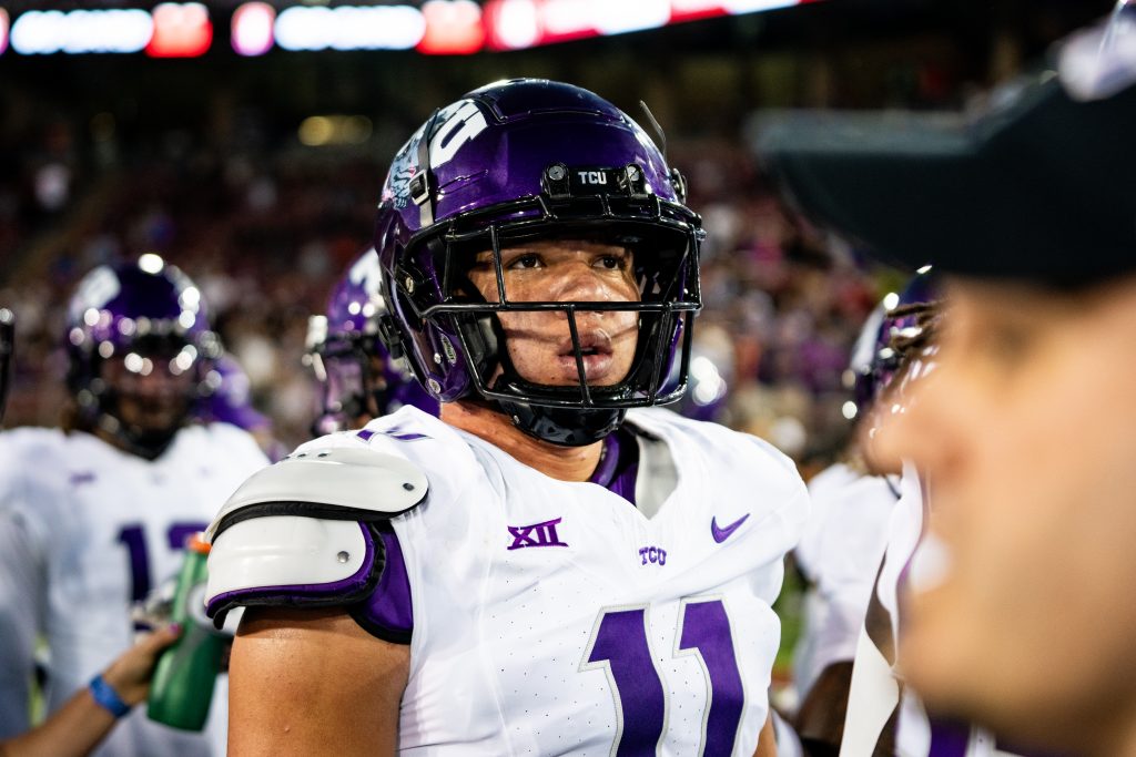 Photograph of TCU football player Devean Deal pictured on the sideline of a football field. Deal is wearing a purple football helmet and white jersey embroidered with a purple number 11. Coaches and teammates of Deal's stand in the image's foreground and background.