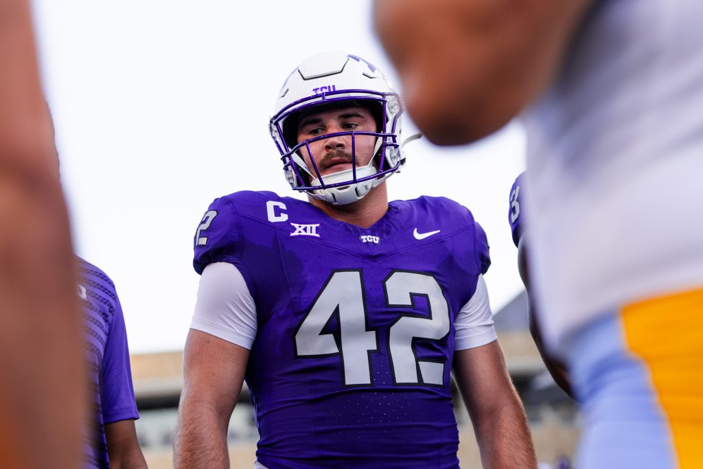 Photograph of TCU long snapper Brent Matiscik wearing a purple football jersey and white helmet. In the foreground, a Long Island University player in a white uniform and powder blue pants frames the right side of the image.