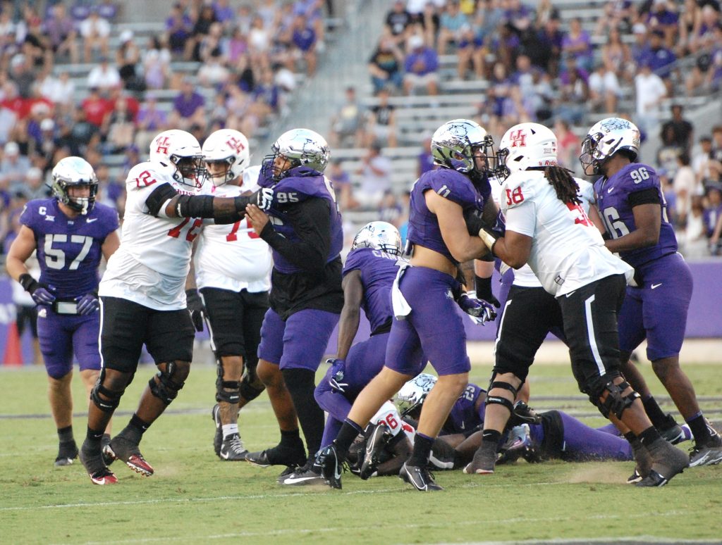 Photograph of football players in white, red and black uniforms from one team and purple and silver uniforms from another team, engaged in a scrum on a grass field, with spectators filling the background.