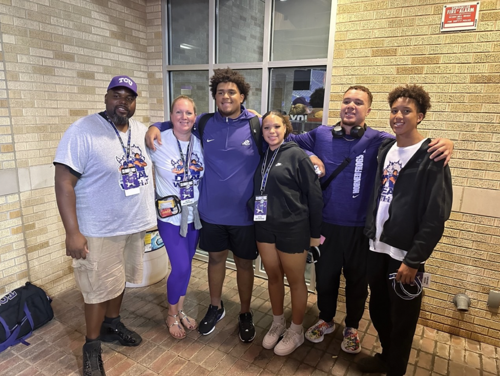 Group photo of six people wearing TCU athletics gear, standing in front of a tan brick wall with a window in the background.