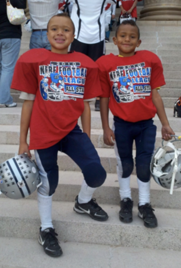 Two individuals in football gear holding silver helmets, standing on stairs, with additional people visible in the upper-left corner of the background.