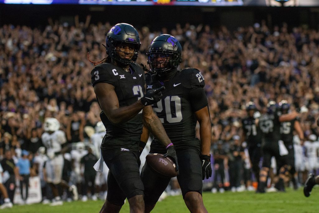 Photograph of TCU wide receiver Savion Williams celebrating a touchdown with teammate Dominique Johnson in a stadium. Both players wear black football helmets, jerseys, and pants. Williams points toward the cheering crowd with his right hand.