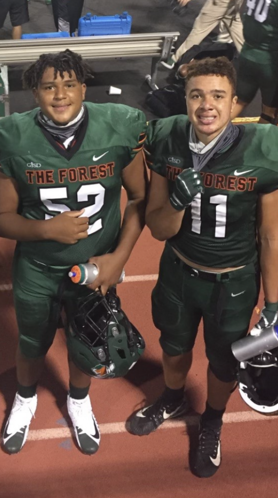 Two individuals wearing green football uniforms stand on a track, smiling at the camera while holding football helmets and water bottles.