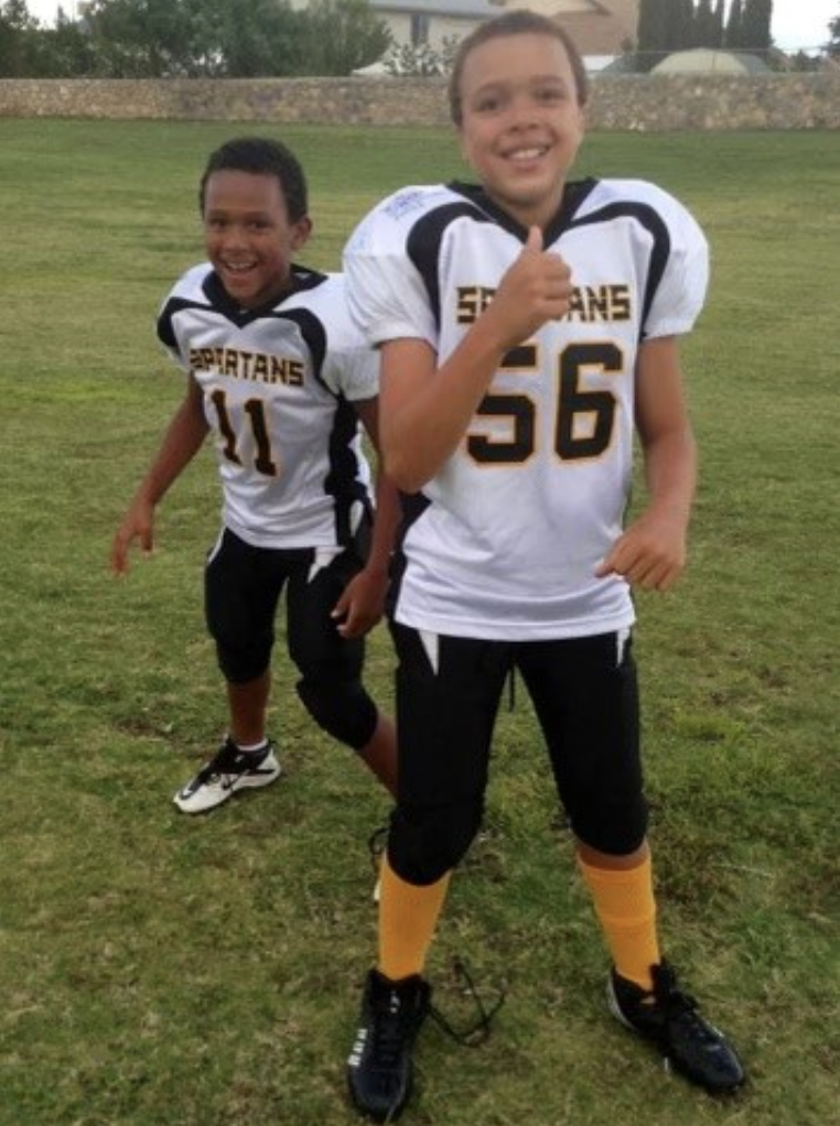 Two individuals dressed in white football jerseys, black football pants and yellow socks smile at the camera while standing on a grass field.