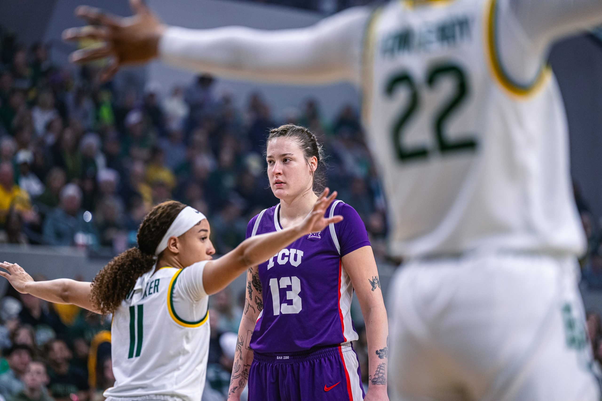 Three basketball players are shown: two in white, green, and gold Baylor women's basketball uniforms, and one in a purple, white, and red TCU uniform. The Baylor players are positioned with their arms outstretched as if defending an inbound pass, while the TCU player, Sedona Prince, looks forward.