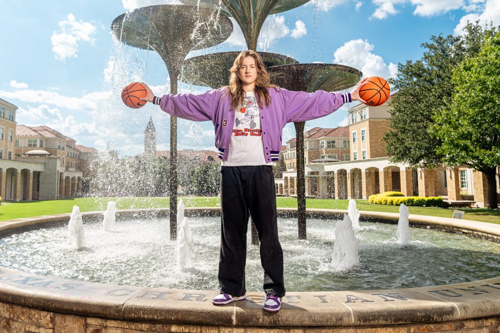 TCU women's basketball player Sedona Prince stands by Frog Fountain on a sunny day, holding a basketball in each hand at shoulder height. She wears a purple jacket, a gray, white, and orange retro TCU basketball shirt, black pants, and white and purple Nike sneakers, looking directly at the camera.