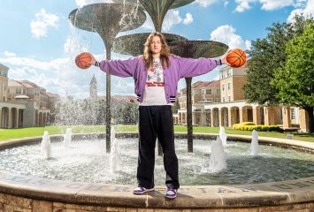 TCU women's basketball player Sedona Prince stands by Frog Fountain on a sunny day, holding a basketball in each hand at shoulder height. She wears a purple jacket, a gray, white, and orange retro TCU basketball shirt, black pants, and white and purple Nike sneakers, looking directly at the camera.