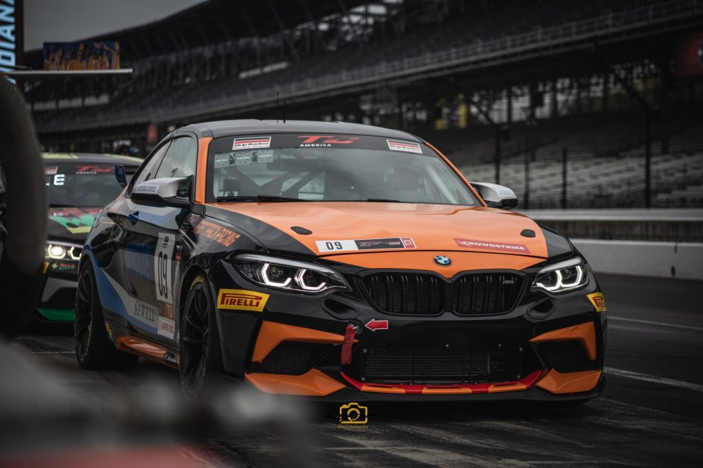 Photograph of an orange and black BMW M6 on a race track on an overcast day. Empty stands fill the image's background.