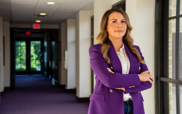 Person in a purple blazer, white dress shirt and blue jeans crosses their arms and looks at the camera, standing in a purple-carpeted hallway.