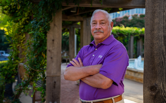 Man in a purple TCU polo shirt crosses his arms and looks at the camera, with vegetation and a water fountain in the background.