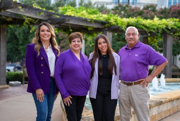 Photograph of four people standing beside a water fountain on Texas Christian University’s campus, with university buildings and landscaping in the background.