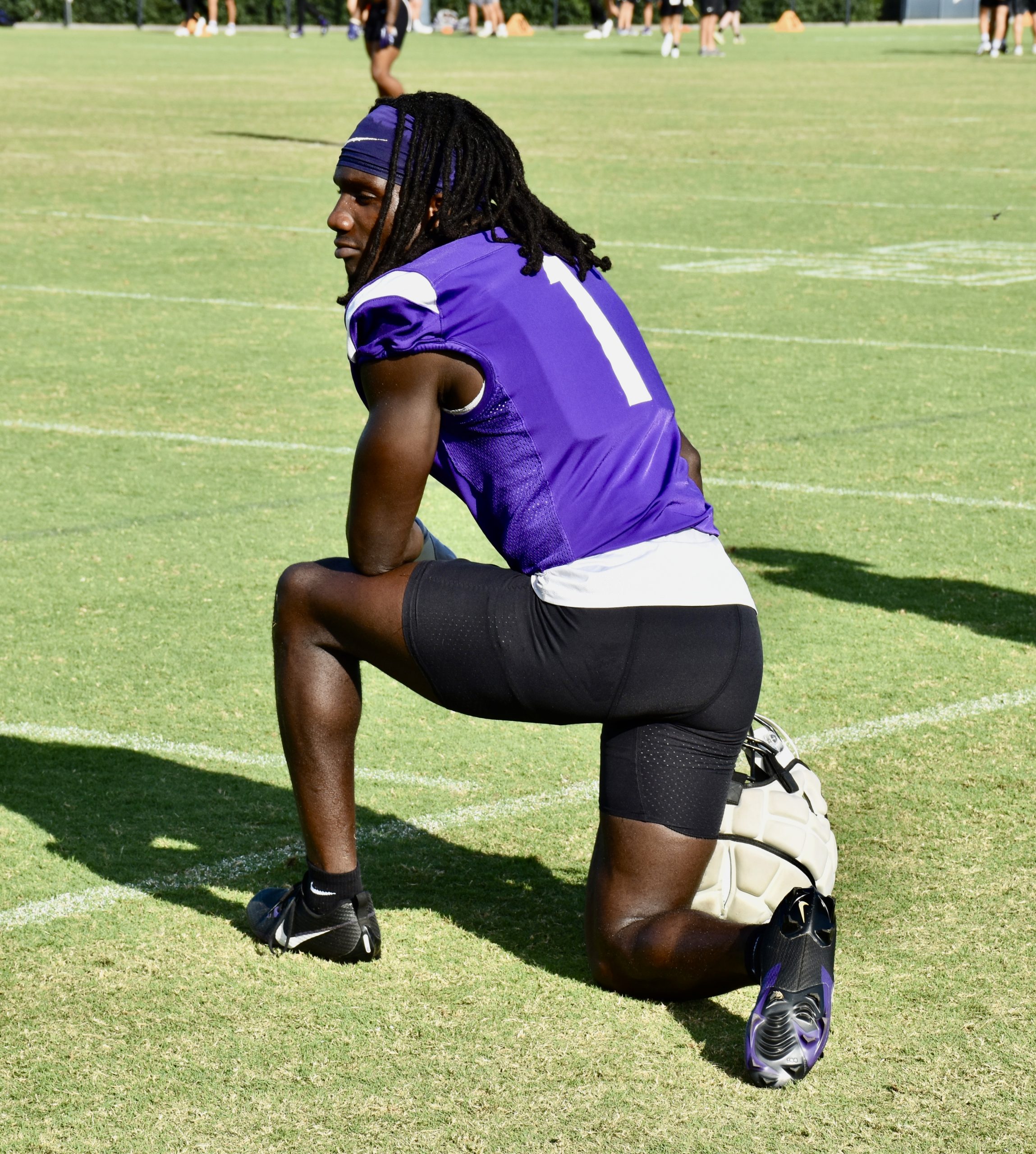 Photograph of TCU football player Abe Camara kneeling on a football practice field, looking to his left. He is wearing a purple practice jersey and black football pants, with the legs of other student-athletes visible in the background.