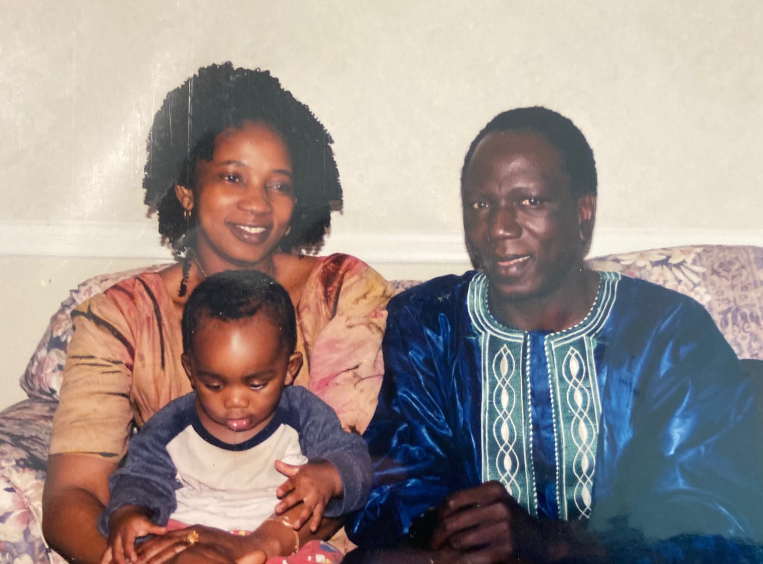 Photograph of two parents smiling while sitting on a flower-print couch, with their infant child on one parent's lap.