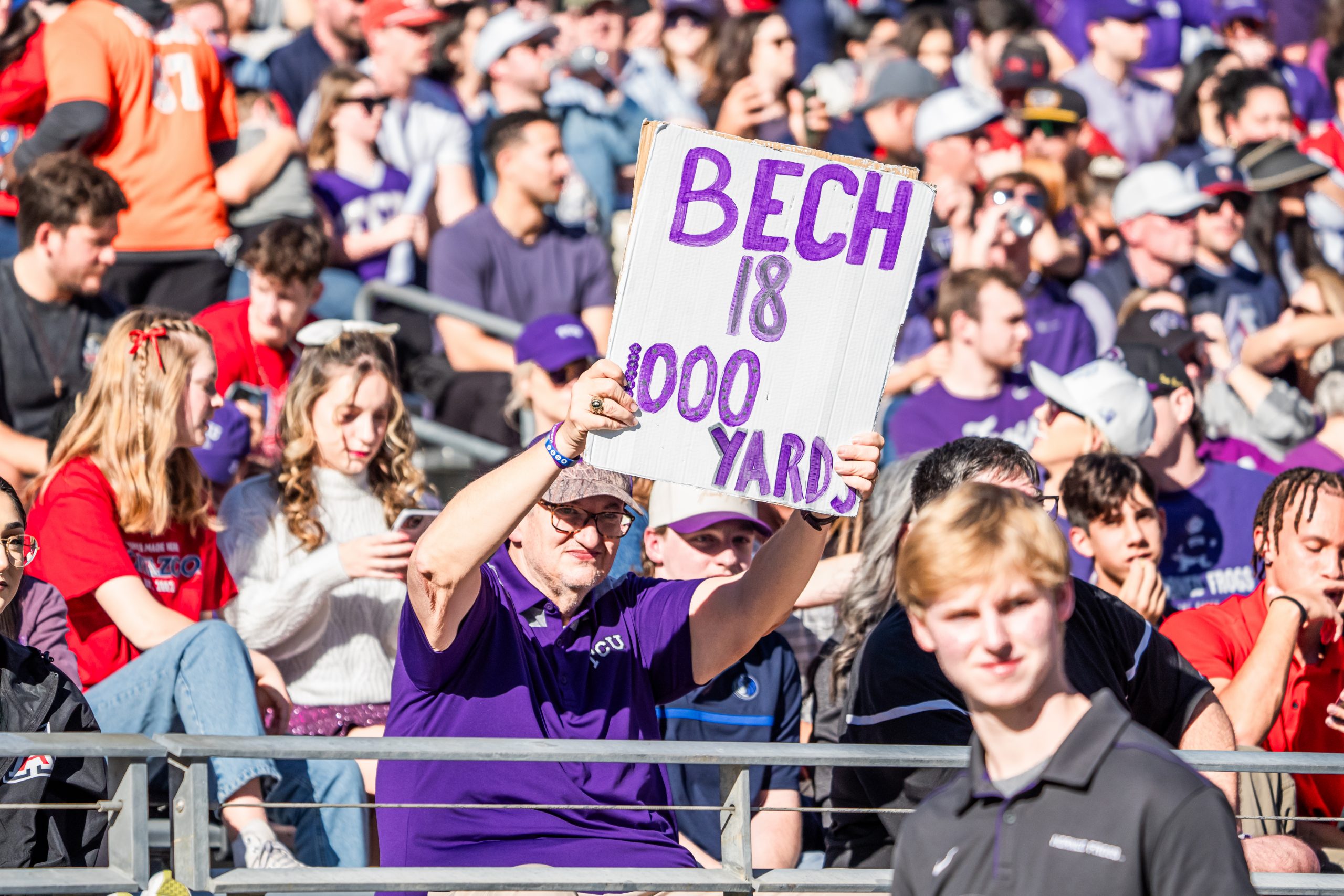 Photograph of a TCU football fan in a purple polo shirt holding up a white sign with purple lettering that reads on three separate lines: "Bech," "18" and "1,000 yards."