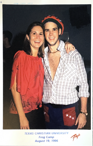 Photograph of two people smiling at the camera in a dimly lit room. The person on the left has their left arm draped over the other's shoulder.