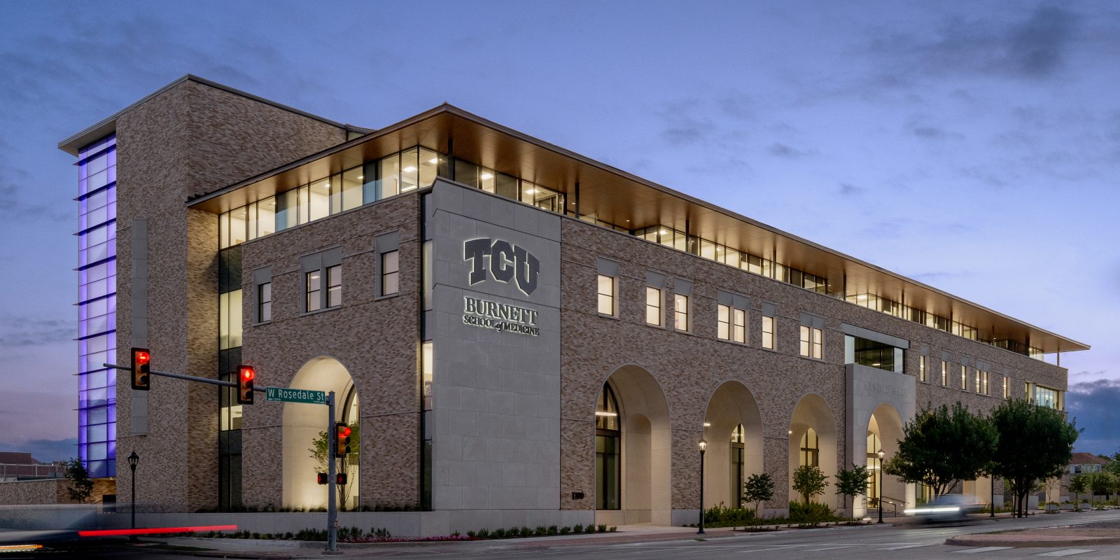 Early morning street-level photo of Arnold Hall, home of The Anne Burnett Marion School of Medicine at Texas Christian University, with a car’s taillights streaked by slow shutter speed.