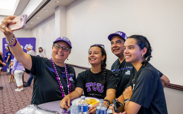 Four people in TCU Horned Frogs apparel take a group selfie in what appears to be a ballroom.