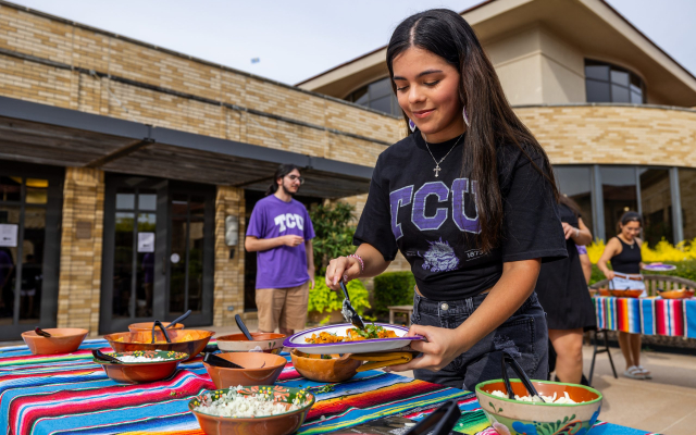 Person in a black TCU t-shirt adds condiments to food on a paper plate, with a brick building and people standing on a patio in the background.