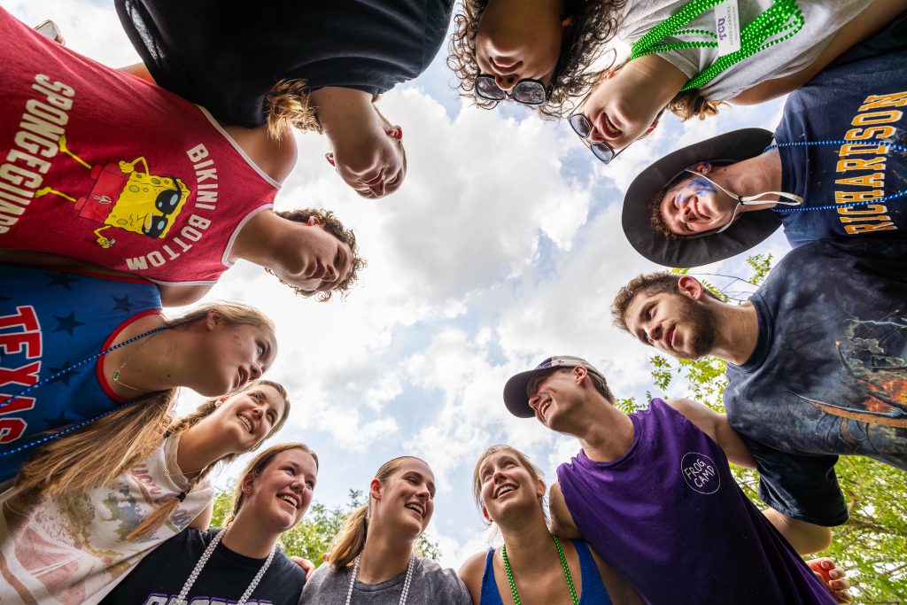 Photograph of a group of students standing in a circle, shoulder-to-shoulder, viewed from below with a blue sky and clouds above.