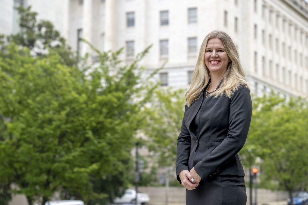 Photograph of a person in a black business suit and dress, standing on a city street with trees and a large building in the background, hands crossed in front.