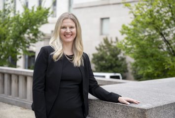 Photograph of a person standing on Capitol Hill in Washington, D.C., wearing a black shirt and black jacket. They are leaning against a concrete railing, smiling toward the camera with a building visible in the background.