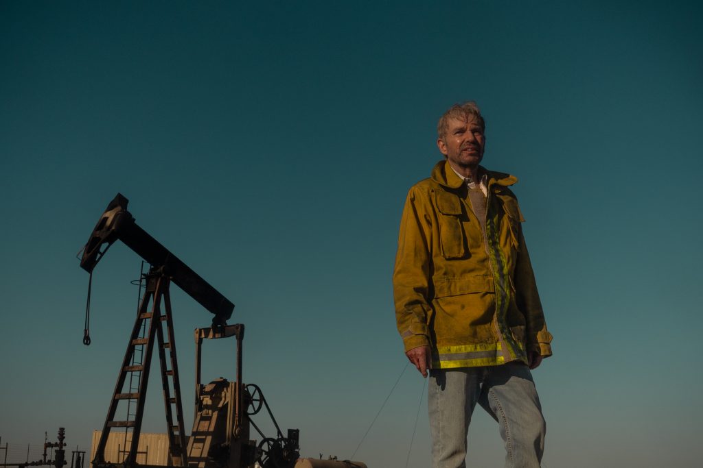 Photograph of Billy Bob Thorton during the filming of the Paramount+ television series “Landman.” Thorton wears a yellow jacket as he stands in front of an oil rig. 