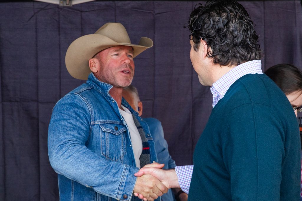 Photograph of two individuals — one in a tannish cowboy hat and jean jacket, the other in a button-down shirt and green-blue sweater — shaking hands.