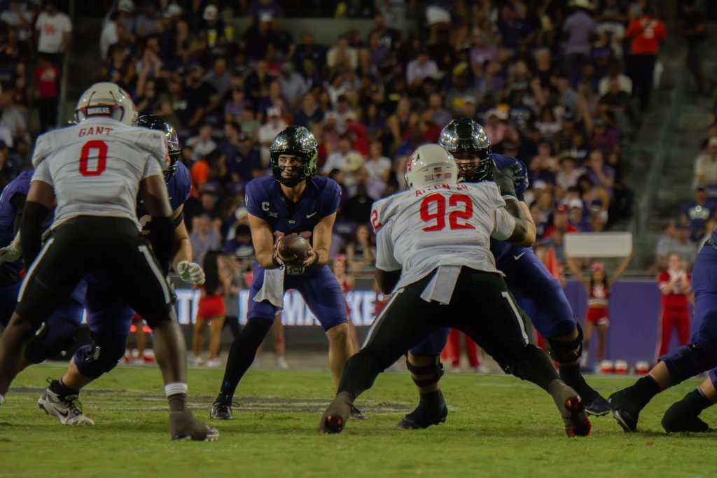 Photograph of TCU quarterback Josh Hoover catching a snap during an October 2024 college football game at Amon G. Carter Stadium in Fort Worth. In front of Hoover, defenders from Houston try to reach the quarterback, while the TCU offensive linemen block.