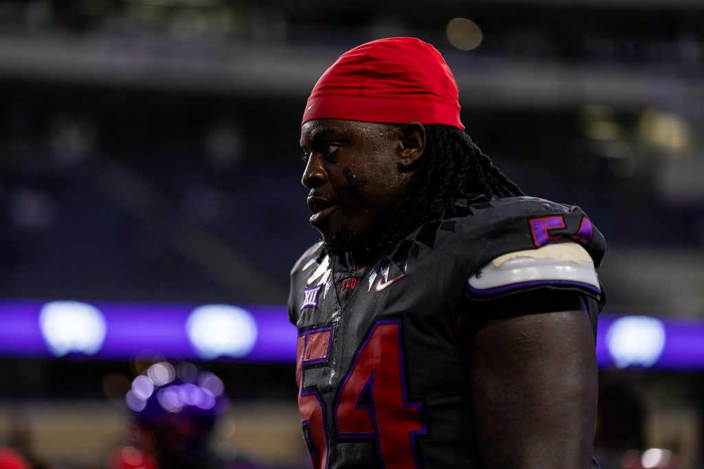 TCU student-athlete NaNa Osafo-Mensah walking on the field at Amon G. Carter Stadium in Fort Worth, Texas, wearing a red skull cap and dark gray TCU football jersey. 