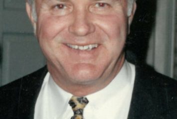 Man in a blazer, white dress shirt, and yellow and blue patterned tie, smiling at the camera.