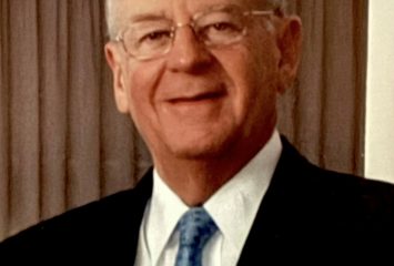 Photograph of a gray-haired person wearing glasses, a teal blue tie, a white dress shirt, and a black sports jacket, smiling at the camera with a brown curtain in the background.