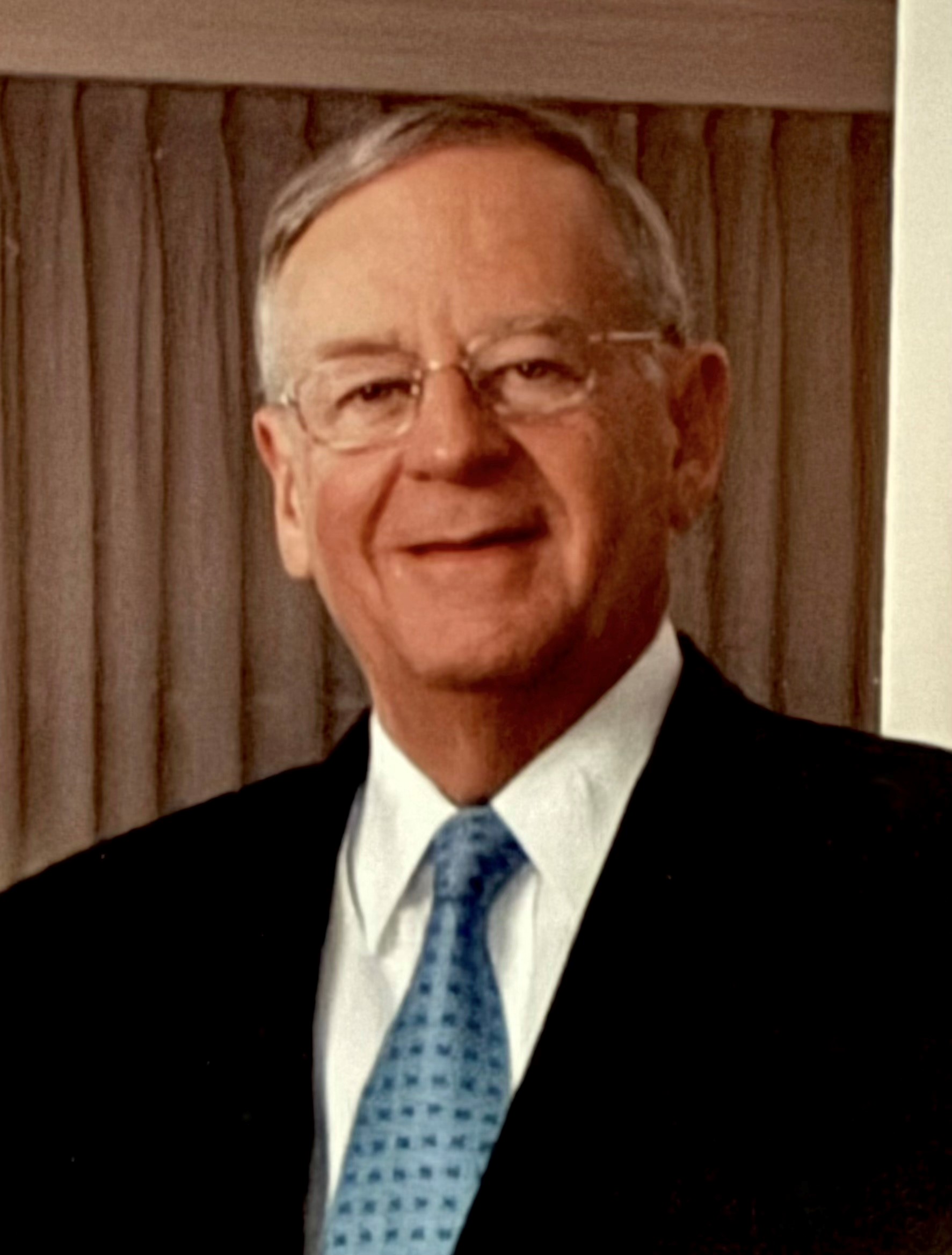 Photograph of a gray-haired person wearing glasses, a teal blue tie, a white dress shirt, and a black sports jacket, smiling at the camera with a brown curtain in the background.