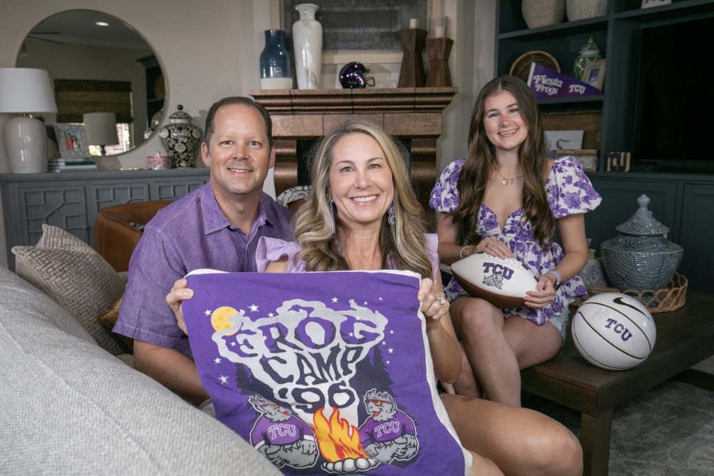 Photograph of three people dressed in purple, seated in a living room and smiling at the camera. The person in front holds up a blanket made of sewn-together t-shirts, featuring text reading “Frog Camp ’96” with two Horned Frogs sitting under a full moon and starry sky beside a campfire. 