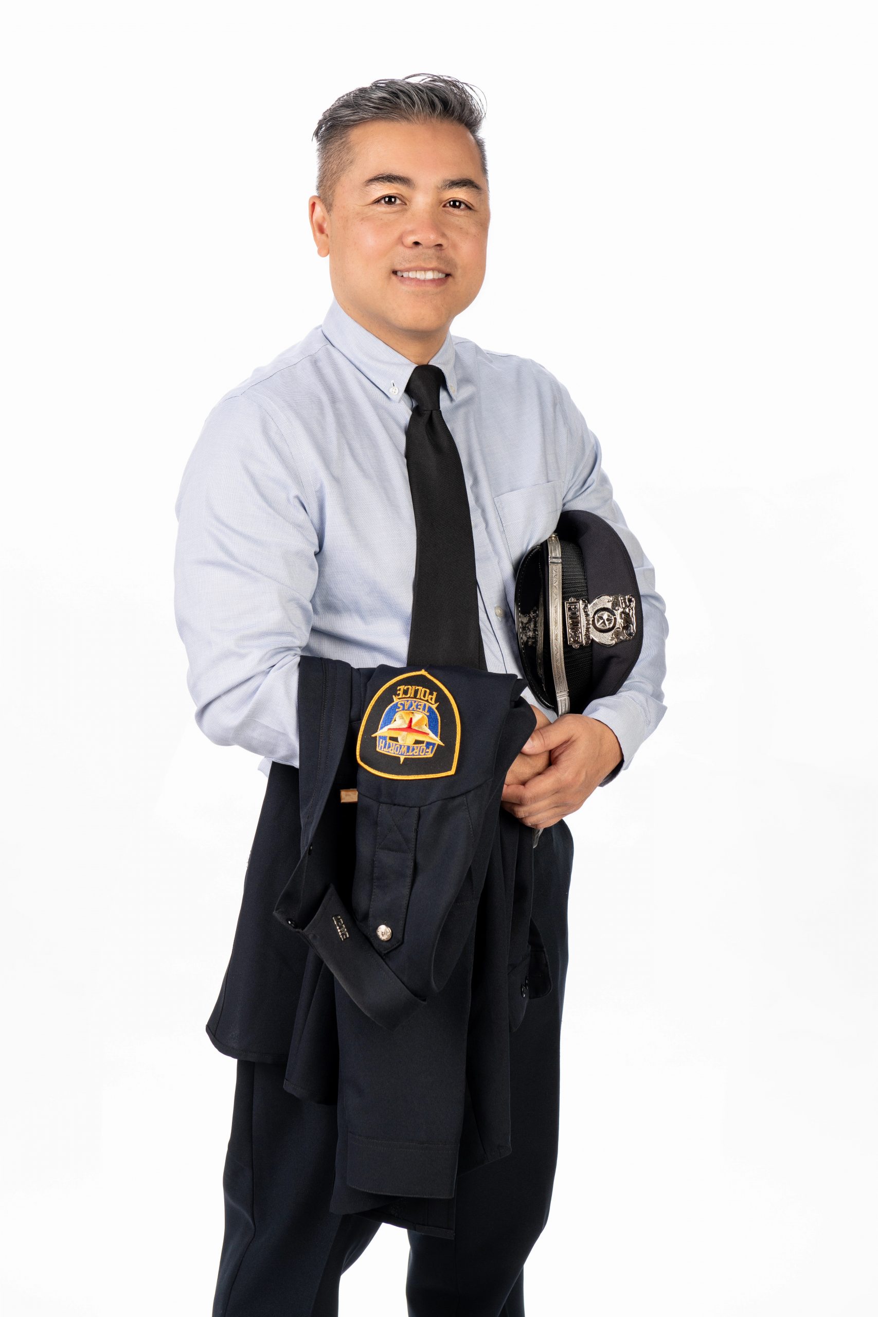 Portrait of a person in a dress shirt and tie, holding a police hat at their side with a calm expression.