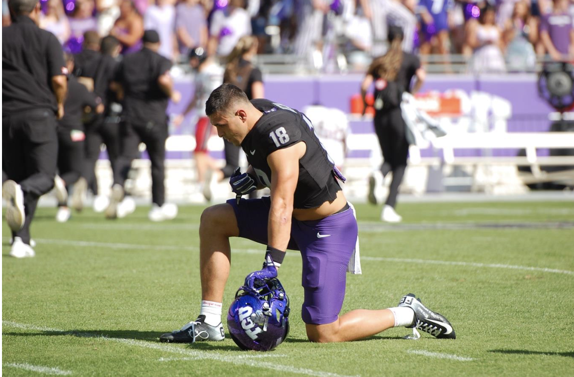 Photograph of TCU football player Jack Bech kneeling in prayer before an October 26, 2024, NCAA football game against Texas Tech. Bech has his face turned downward toward the field, his right hand hovering over his chest and his left resting on his purple TCU football helmet.