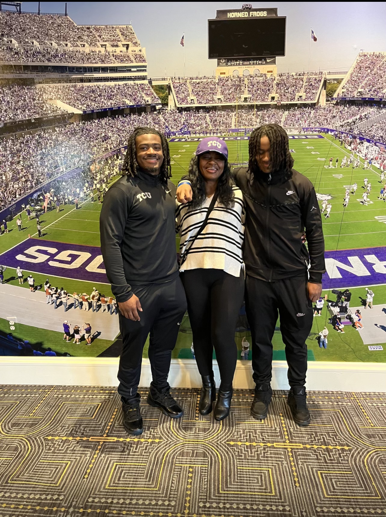 Photograph of three people standing on a yellow, silver and black carpeted floor, smiling toward the camera, as they pose in front of a wall-length photograph of Amon G. Carter Stadium on a sunny game day. 