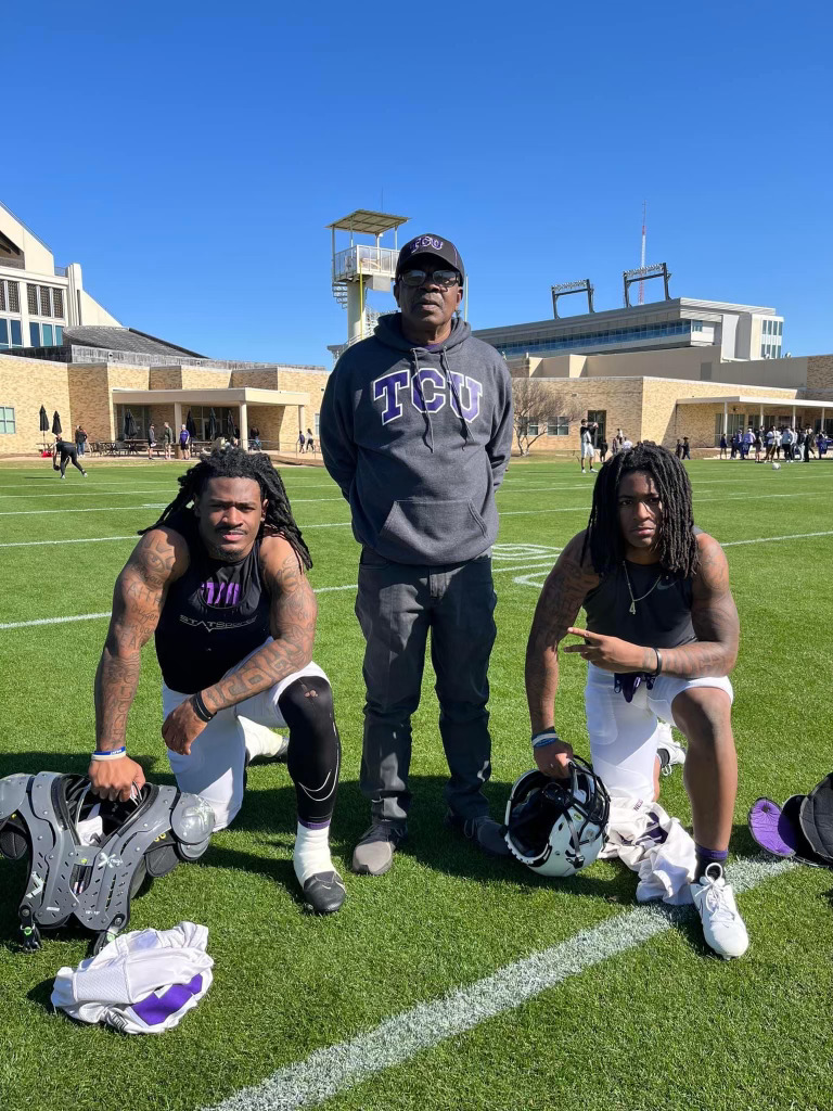 Two individuals wearing black athletic t-shirts and white football pants kneel on a football practice field. The person on the left places their right hand on a pair of shoulder pads, while the person on the right rests their right hand on a football helmet. Between them stands an older individual wearing a grey TCU sweatshirt.