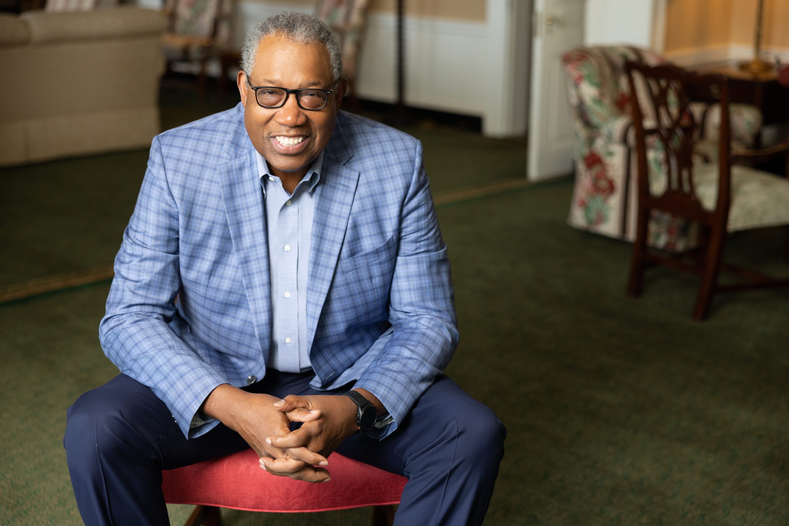 Photograph of a smiling James Cash in a light blue dress shirt, blue blazer and navy pants, leaning forward with his hands crossed in front of him in a cardinal red, patterned chair.