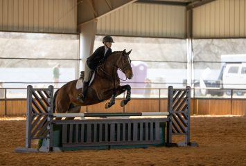 Photograph of TCU equestrian team member Ashleigh Scully on horseback, leaping over a fence during an NCAA meet against Baylor in March 2023. Scully is looking straight ahead.