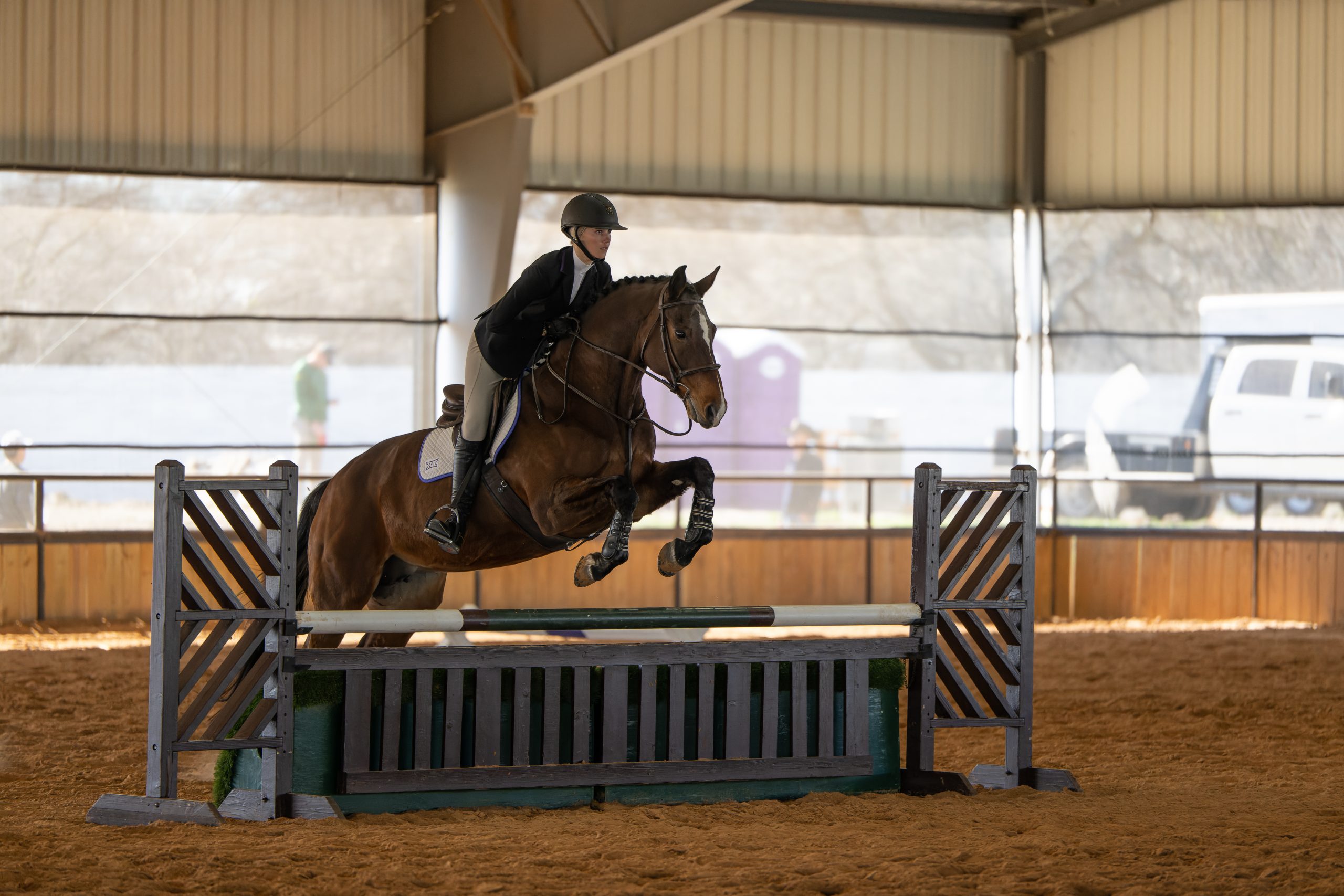 Photograph of TCU equestrian team member Ashleigh Scully on horseback, leaping over a fence during an NCAA meet against Baylor in March 2023. Scully is looking straight ahead.