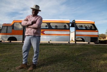 Artist and writer Christopher Blay stands in a grassy area with arms crossed in front of his art installation, a repurposed 1970s-era bus shell.