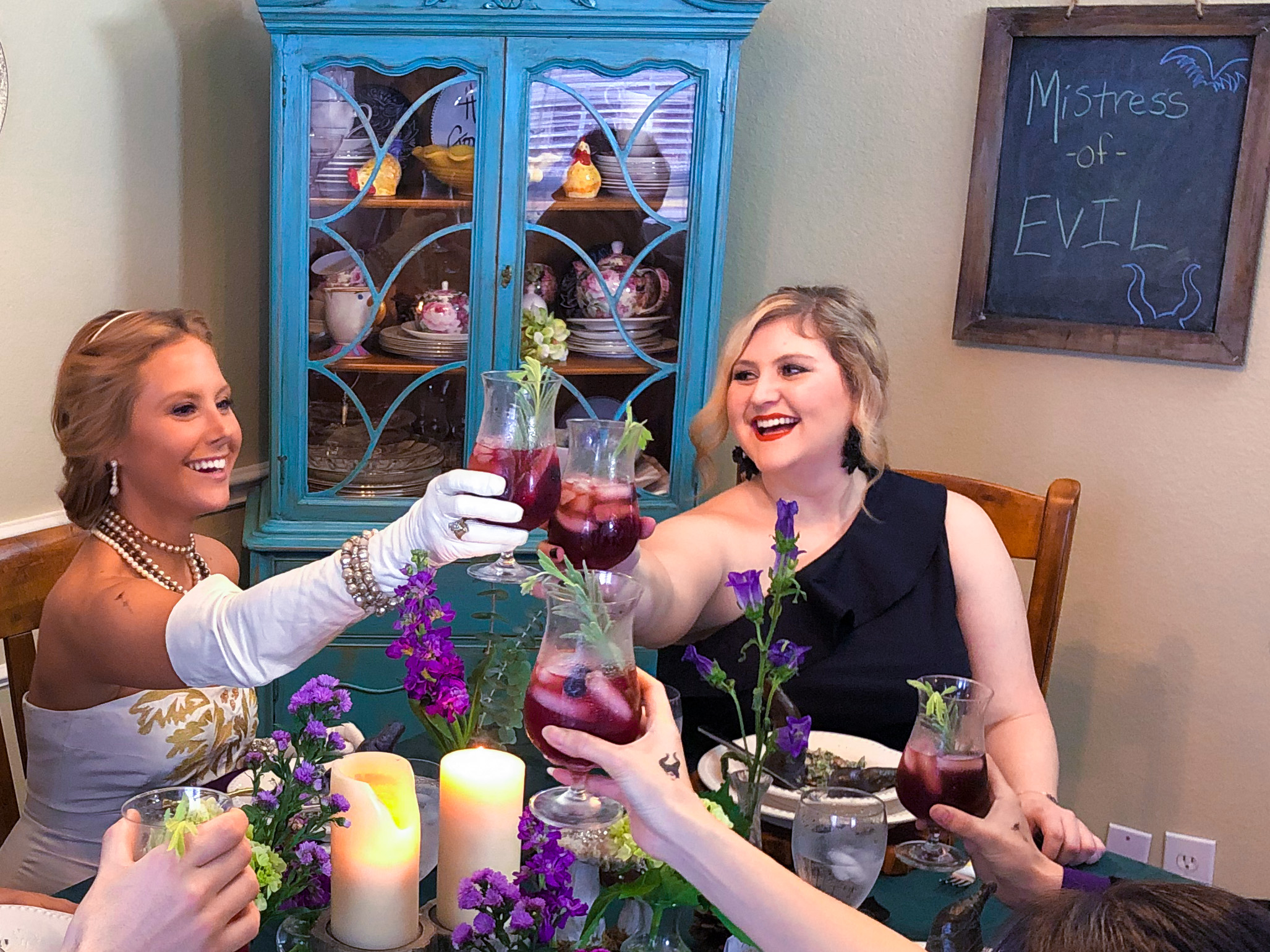 Katie-Rose Watson sits at a dining room table, smiling as she clinks glasses with others. One attendee wears a Disney princess-style white glove.