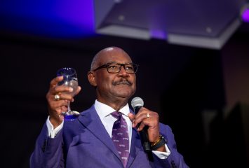 Ronald Parker holds a microphone near his mouth in his left hand and a glass of water in his right, appearing to make a toast.