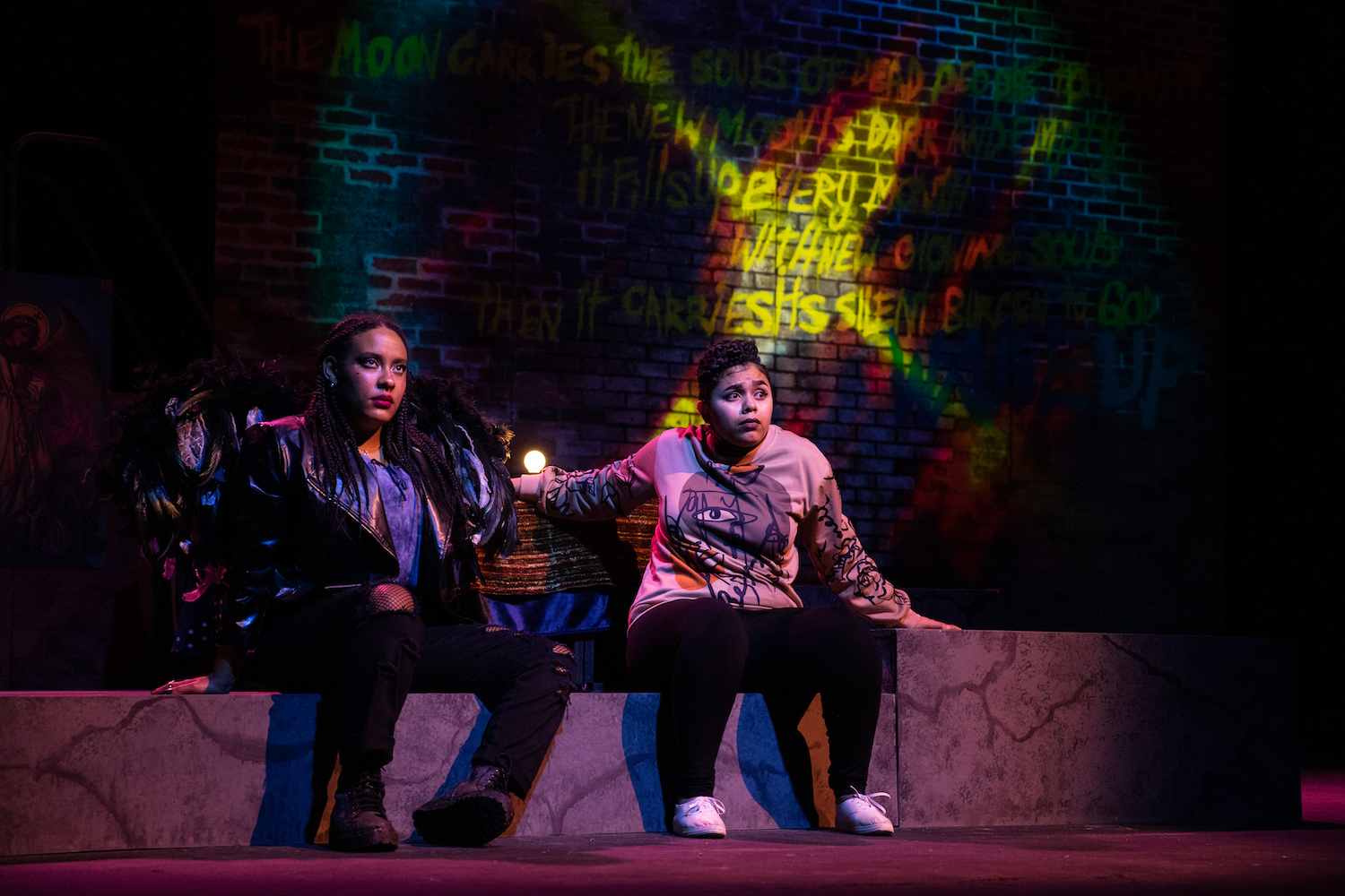 Photograph of two student actors sitting side by side on a ledge or bench, looking straight ahead. Behind them, brightly illuminated words in greens, yellows, reds, and blues cover a brick wall, glowing with varying intensity.