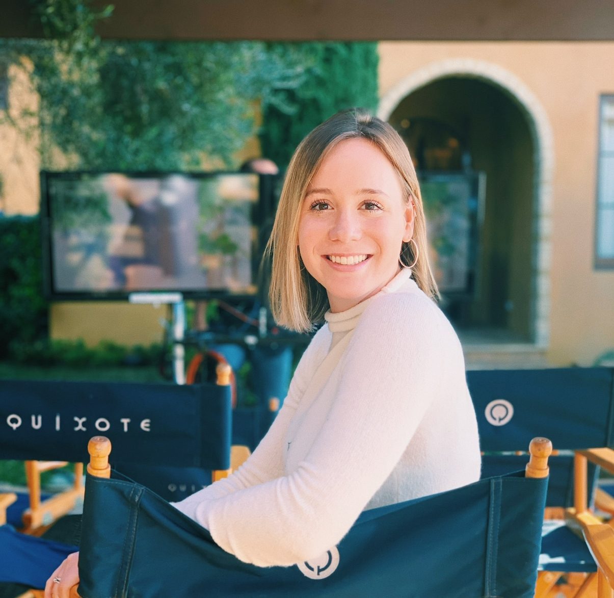 Photograph of Miranda Speir, smiling as she sits in a director’s chair. A filming set fills out the background.