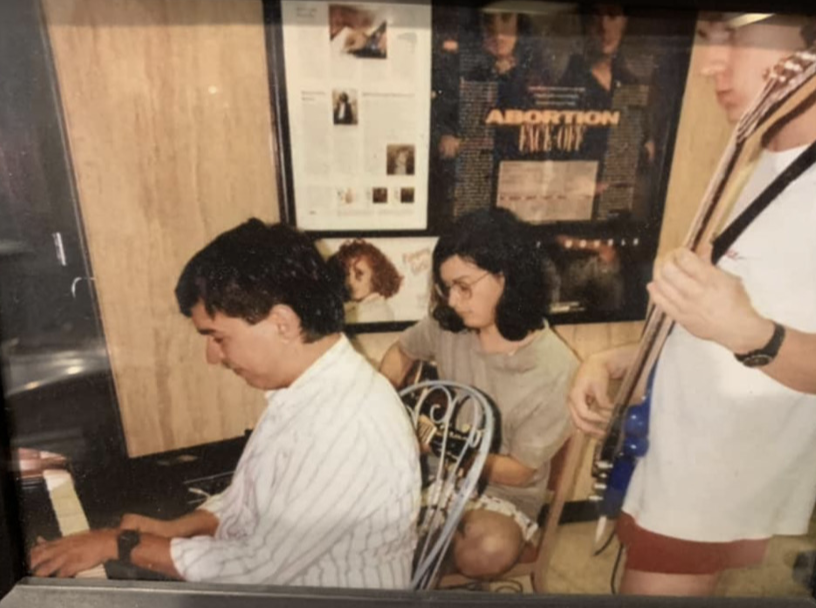 Photograph of three college students seated in a dorm room, playing instruments — two on guitar or bass, one on piano.
