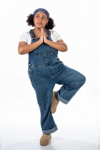Photograph of Theatre TCU student Jess Willis balancing on one leg, hands clasped over her chest, gazing to the right against an all-white background.